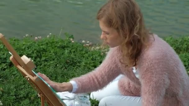 Красивая женщина с рыжими волосами, рисует картину на холсте, который стоит на мольберте. The lady is in the open air near the lake of the river, she draw from life — стоковое видео