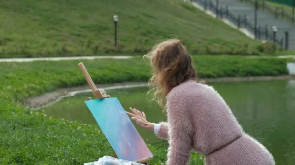 Eine hübsche Frau mit roten Haaren malt ein Bild auf Leinwand, das auf der Staffelei steht. die Dame steht an der frischen Luft am Ufer des Flusses, sie schöpft aus dem Leben — Stockvideo