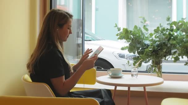 Üzleti nő ül egy asztalnál egy kávézóban kávét iszik, és működő-ra egy tabletta — Stock videók