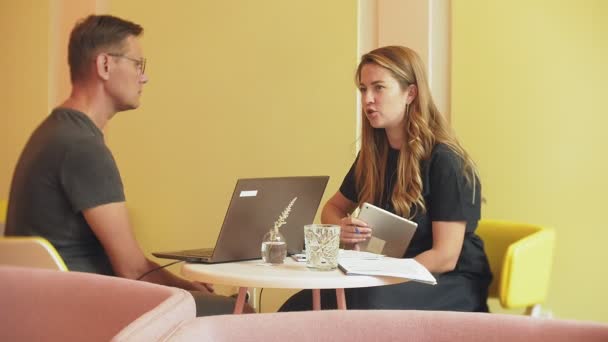 一个商业妇女和一个男人在他们的休息与他们的平板电脑坐在一个黄色的墙壁咖啡馆和讨论项目4K — 图库视频影像