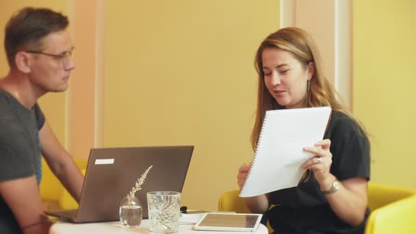 一个商业妇女和一个男人在他们的休息与他们的平板电脑坐在一个黄色的墙壁咖啡馆和讨论项目 — 图库视频影像
