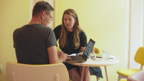 Una mujer de negocios y un hombre en su descanso con su tableta se sientan en un café con paredes amarillas y discuten un proyecto — Vídeos de Stock