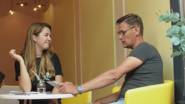 Бізнес-леді і чоловік на перерві зі своїм планшетом сидять в кафе з жовтими стінами і обговорюють проект — стокове відео
