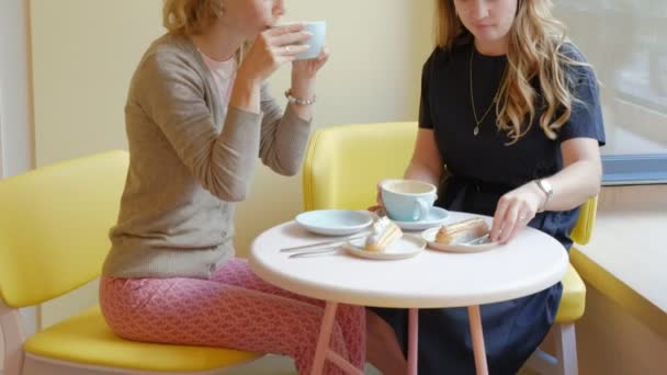 Две женщины, сидящие в кафе-ресторане, едят пирожные эклер, пьют кофе и смеются. — стоковое видео