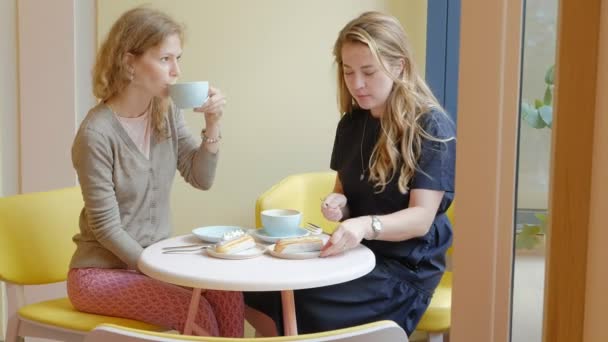 Dos mujeres sentadas en un restaurante cafetería comiendo pasteles eclair bebiendo café y riendo — Vídeo de stock