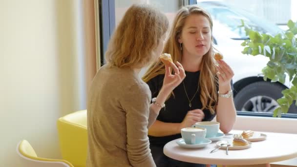 Dos mujeres sentadas en un restaurante cafetería comiendo pasteles eclair bebiendo café y riendo — Vídeo de stock