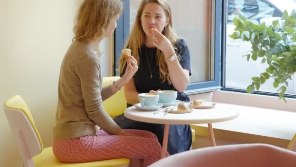 Две женщины, сидящие в кафе-ресторане, едят пирожные эклер, пьют кофе и смеются. — стоковое видео