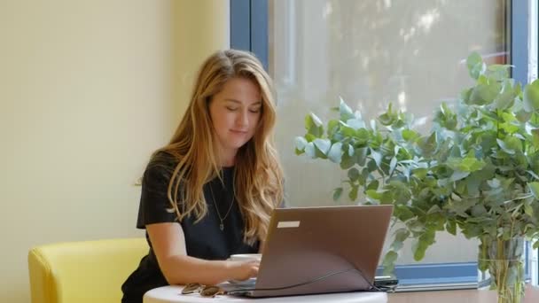 Mujer de negocios sentada en una mesa en un café tomando café y trabajando en un portátil — Vídeos de Stock