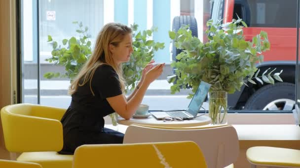 Επιχειρήσεων γυναίκα κάθεται σε ένα τραπέζι σε ένα καφέ, πίνοντας καφέ και να εργάζονται σε ένα φορητό υπολογιστή — Αρχείο Βίντεο
