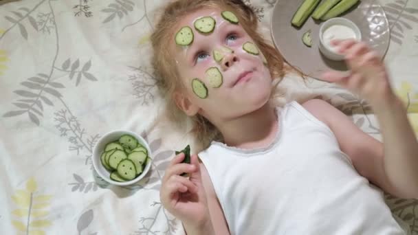 Pequena bela menina bonita está fazendo máscaras de pepino em casa e comer legumes — Vídeo de Stock