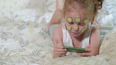 Küçük bir güzel kız neşeyle bir salatalık maskesi evde yapar ve bir akıllı telefon kullanır