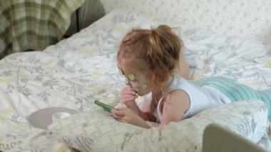 Küçük bir güzel kız neşeyle bir salatalık maskesi evde yapar ve bir akıllı telefon kullanır