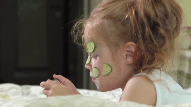 Küçük bir güzel kız neşeyle bir salatalık maskesi evde yapar ve bir akıllı telefon kullanır