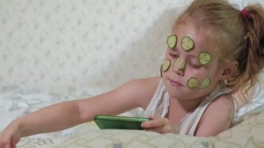 Küçük bir güzel kız neşeyle bir salatalık maskesi evde yapar ve bir akıllı telefon kullanır