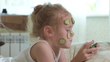 Küçük bir güzel kız neşeyle bir salatalık maskesi evde yapar ve bir akıllı telefon kullanır