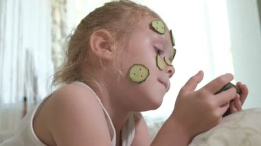 Küçük bir güzel kız neşeyle bir salatalık maskesi evde yapar ve bir akıllı telefon kullanır