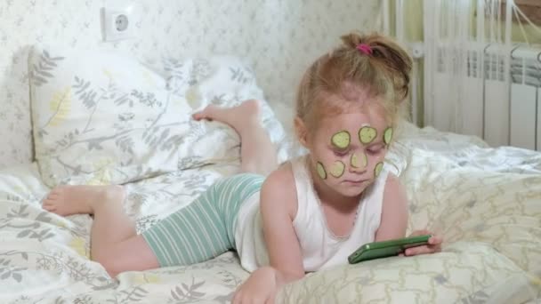 Ένα πανέμορφο κοριτσάκι χαρωπά κάνει ένα αγγούρι μάσκες στο σπίτι και χρησιμοποιεί ένα smartphone — Αρχείο Βίντεο