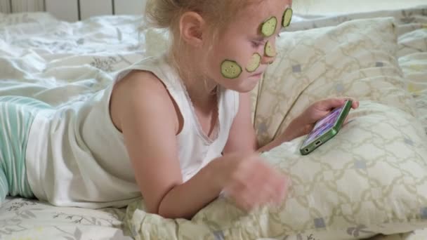 Een mooi meisje vrolijk maakt een komkommer maskers thuis en maakt gebruik van een smartphone — Stockvideo