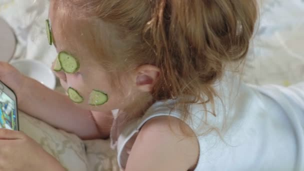 Una niña hermosa alegre hace una máscara de pepino en casa y utiliza un teléfono inteligente — Vídeo de stock