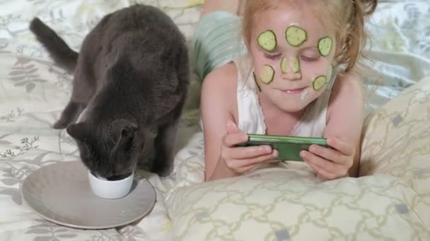 Egy gyönyörű kislány vidáman teszi egy uborkát maszkok otthon, és élvezi egy okostelefon, a macska sits közeli — Stock videók