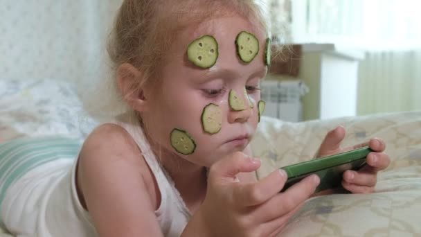 En liten vacker flicka glatt gör en gurka masker hemma och använder en smartphone — Stockvideo