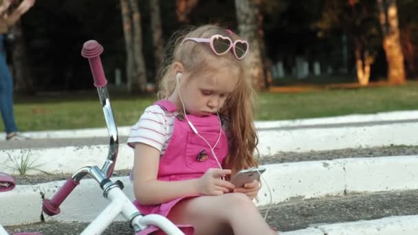Una niña hermosa en un vestido rosa está sentada en el parque en las escaleras y disfruta de un teléfono inteligente en los auriculares, la bicicleta está acostada una al lado de la otra — Vídeo de stock
