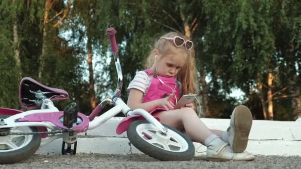 Маленькая красивая девочка в розовом платье сидит в парке на ступеньках и наслаждается смартфоном в наушниках, велосипед лежит рядом друг с другом — стоковое видео