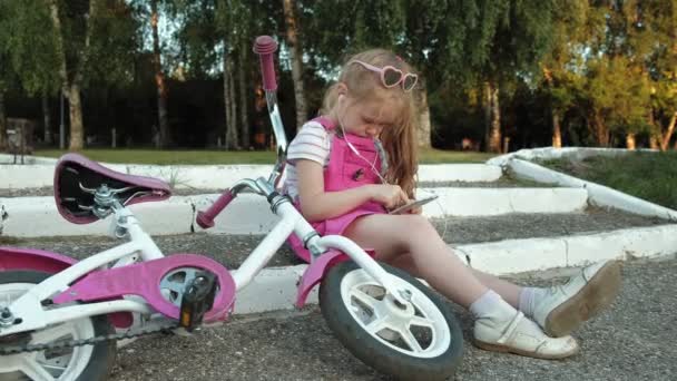 隣同士に自転車が横になっているピンクのドレスを着た美しい少女はステップの上の公園に座っているとヘッドフォンでスマート フォンを楽しんでいます、 — ストック動画