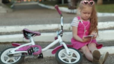 Pembe elbiseli küçük bir güzel kız parkta adımlar üzerinde oturan ve bir akıllı telefon kulaklık sahiptir, Bisiklet yan yana yatıyor