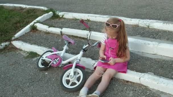 Trochę piękna dziewczyna w różowej sukience siedzi w parku na schodach i cieszy się smartfon w słuchawkach, rower jest leżące obok siebie — Wideo stockowe