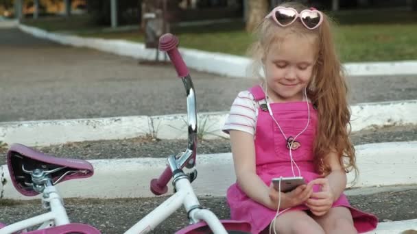 一个穿着粉红色连衣裙的小漂亮女孩坐在台阶上的公园里, 享受着耳机里的智能手机, 自行车就在彼此的旁边。 — 图库视频影像