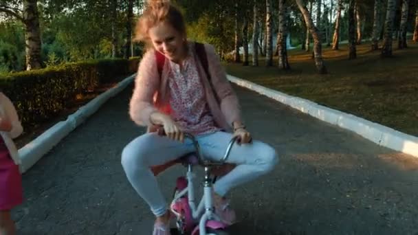 Una niña feliz, hermosa, con el pelo largo y rubio en una falda rosa y saltador monta una bicicleta para niños en la carretera, sonríe . — Vídeos de Stock