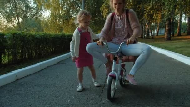 Una niña feliz, hermosa, con el pelo largo y rubio en una falda rosa y saltador monta una bicicleta para niños en la carretera, sonríe . — Vídeo de stock