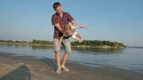Close-up van vader en dochter samenspelen. Een jonge vader draait zijn dochter op het strand van de zee oever super slow motion — Stockvideo