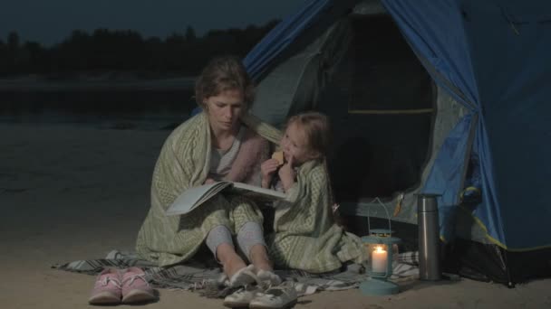 母親と彼女の美しい娘はビーチで夜観光テント近く本を読む — ストック動画