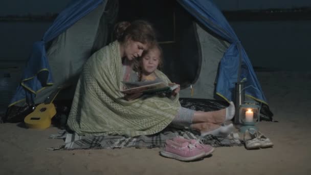 Mutter und ihre schöne Tochter lesen nachts am Strand in der Nähe eines Touristenzeltes ein Buch — Stockvideo