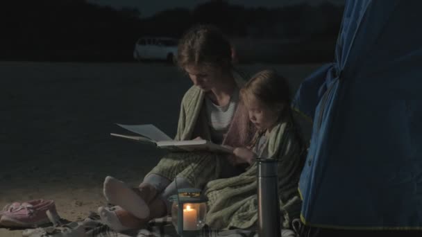 Madre e la sua bellissima figlia leggono un libro vicino a una tenda turistica di notte sulla spiaggia — Video Stock