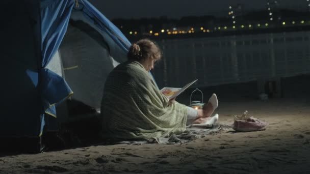 Madre e la sua bellissima figlia leggono un libro vicino a una tenda turistica di notte sulla spiaggia — Video Stock