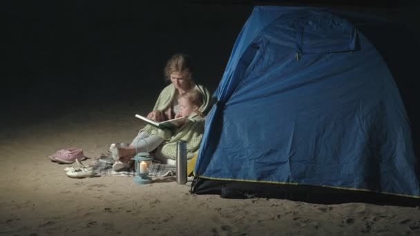 Mãe e sua linda filha lêem um livro perto de uma tenda turística à noite na praia — Vídeo de Stock