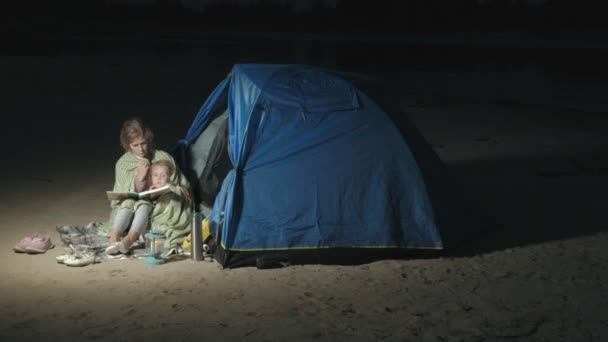 Anya és lánya szép olvasni egy könyvet, közel a turisztikai sátor éjjel a parton — Stock videók