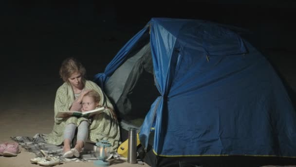 Mor och hennes vackra dotter läsa en bok nära turist tält på natten på stranden — Stockvideo