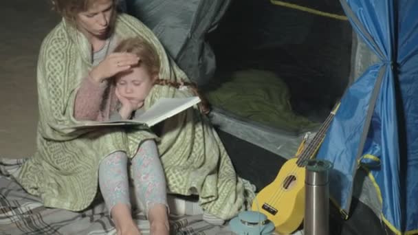 母亲和她美丽的女儿在海滩上晚上在旅游帐篷附近看书。 — 图库视频影像