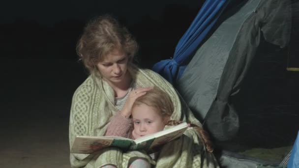 Madre y su hermosa hija leen un libro cerca de una tienda de campaña turística por la noche en la playa — Vídeo de stock