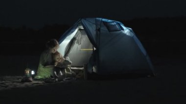 Anne ve onun güzel kızı gece sahilde bir turist çadır yakınındaki bir kitap okumak