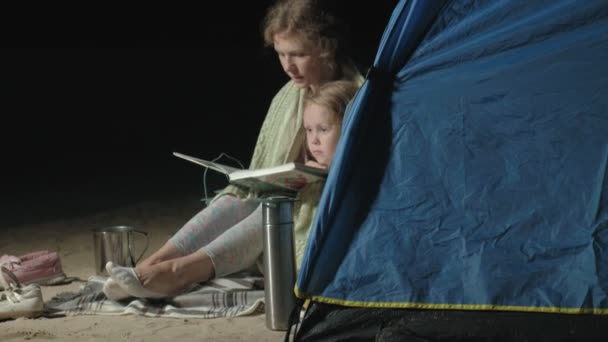 Mutter und ihre schöne Tochter lesen nachts am Strand in der Nähe eines Touristenzeltes ein Buch — Stockvideo