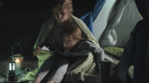 Mãe e sua linda filha lêem um livro perto de uma tenda turística à noite na praia — Vídeo de Stock