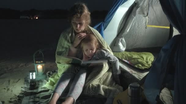 Madre y su hermosa hija leen un libro cerca de una tienda de campaña turística por la noche en la playa — Vídeos de Stock
