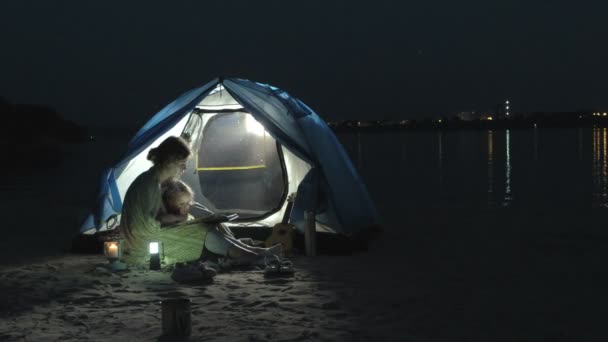 Mãe e sua linda filha lêem um livro perto de uma tenda turística à noite na praia — Vídeo de Stock