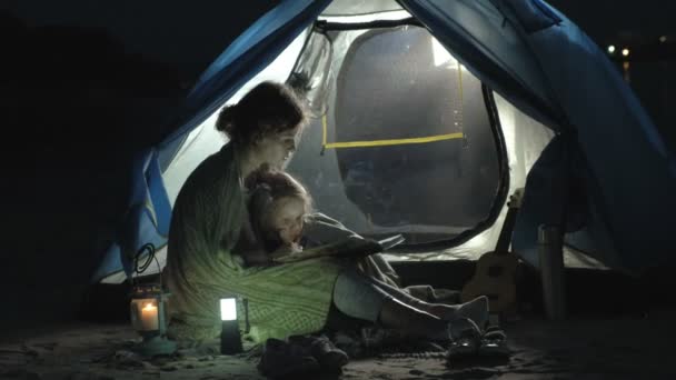 Madre e la sua bellissima figlia leggono un libro vicino a una tenda turistica di notte sulla spiaggia — Video Stock