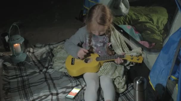 En vacker flicka spelar på en ukulele på flodstranden nära turist tält — Stockvideo